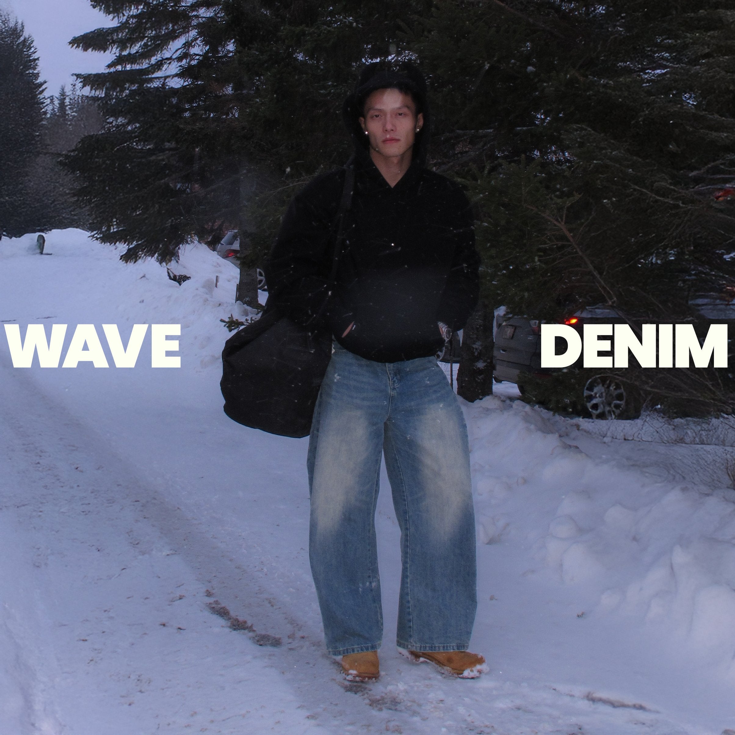 Person wearing wide-leg denim jeans and black hoodie on snowy forest road, Wave Denim text