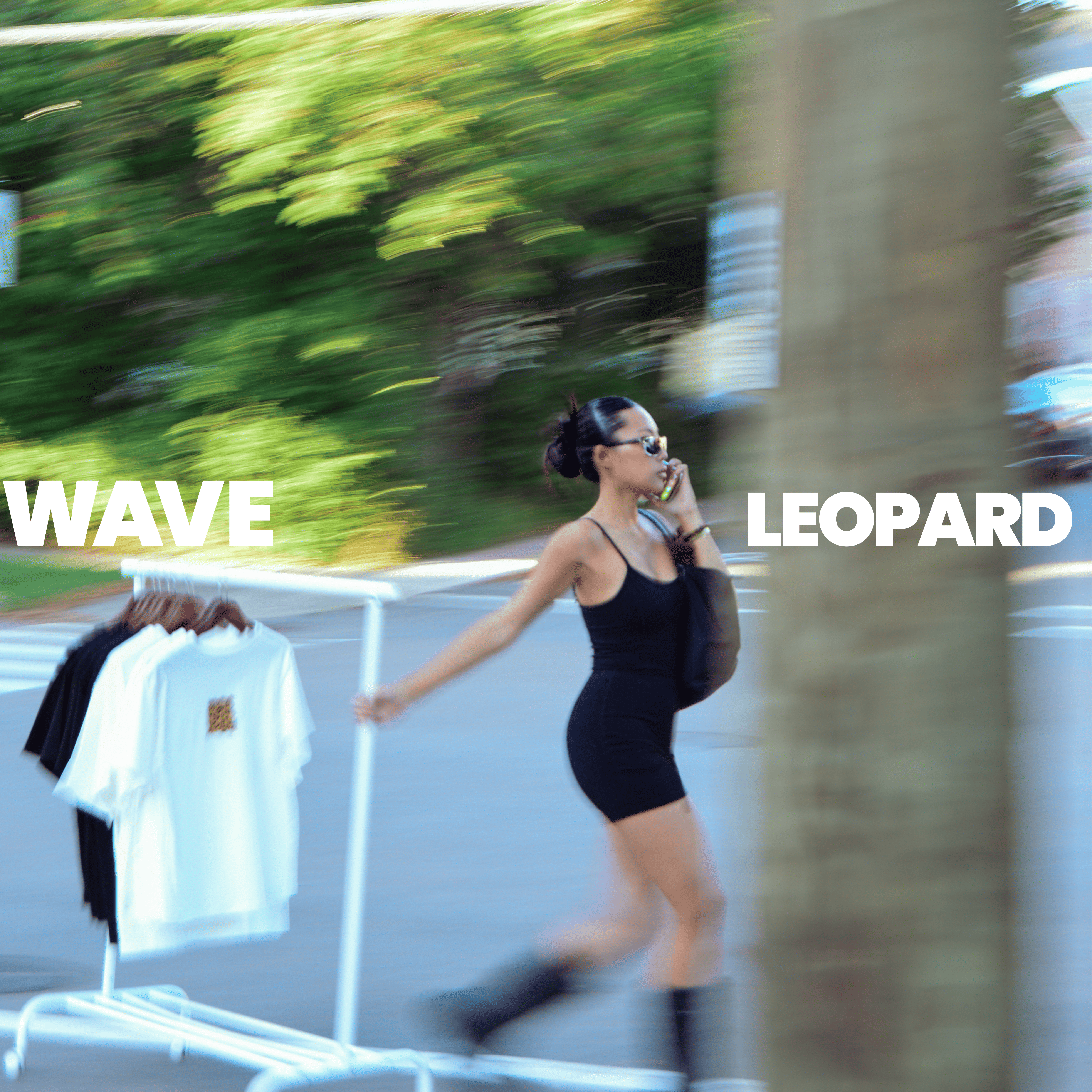 Woman in black dress walking past rack of graphic t-shirts outdoors, text 'WAVE LEOPARD'
