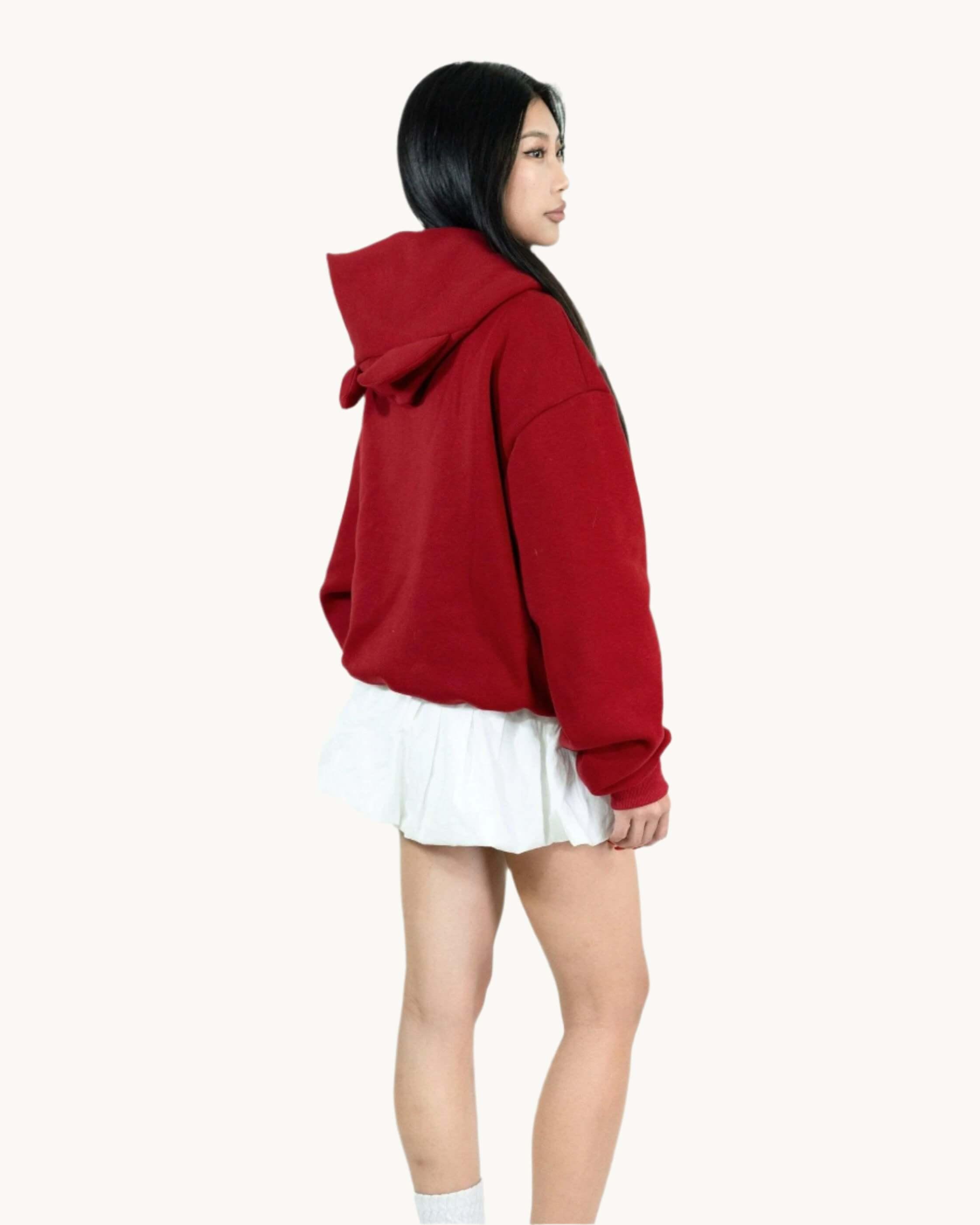 Woman in red hoodie and white skirt standing against plain background, side profile view